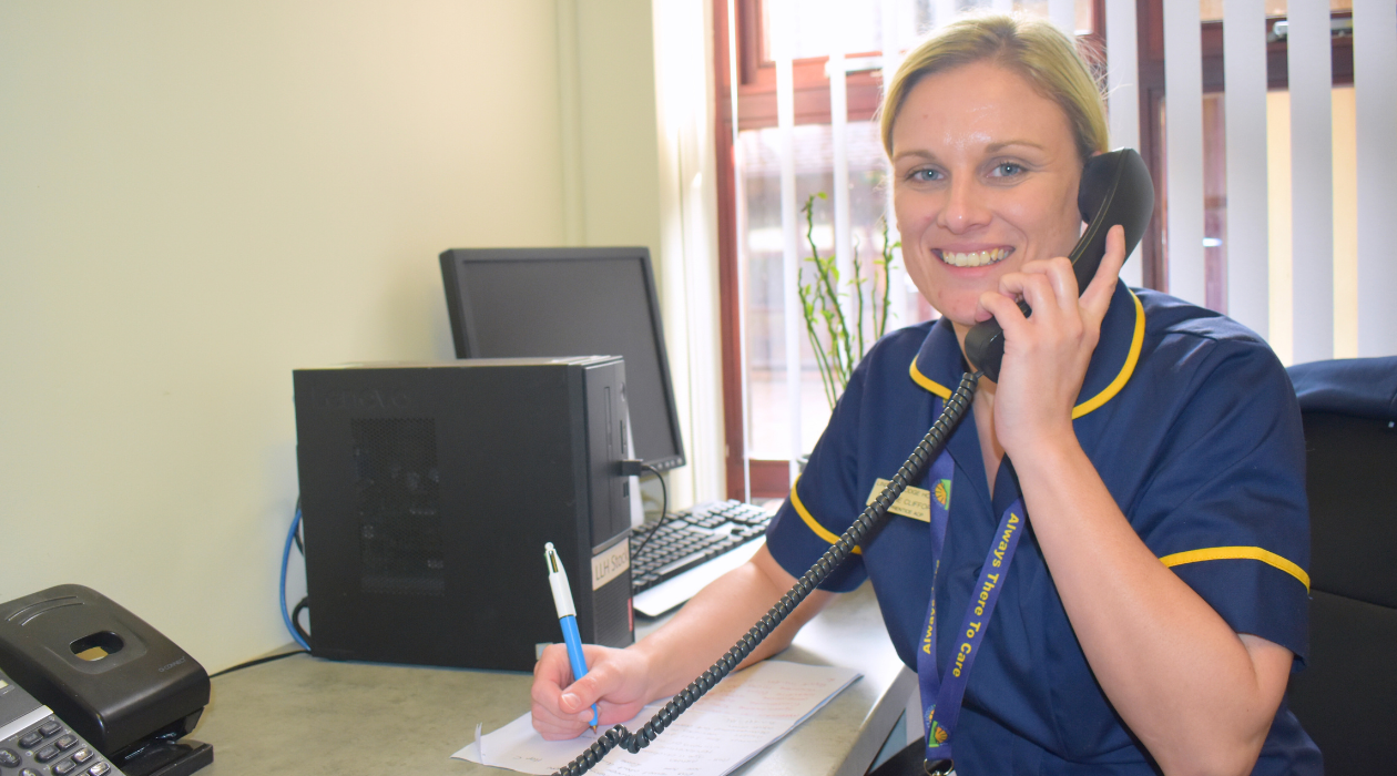 Image of Advanced care practitioner Sophie on the phone. Sophie is dress in a dark blue nurses uniform with a yellow trim, with blonde hair and a smile on her face. She is holding a black landline phone to her ear and a blue and white pen in her other hand, which is resting on a piece of paper on the desk ready to take notes.